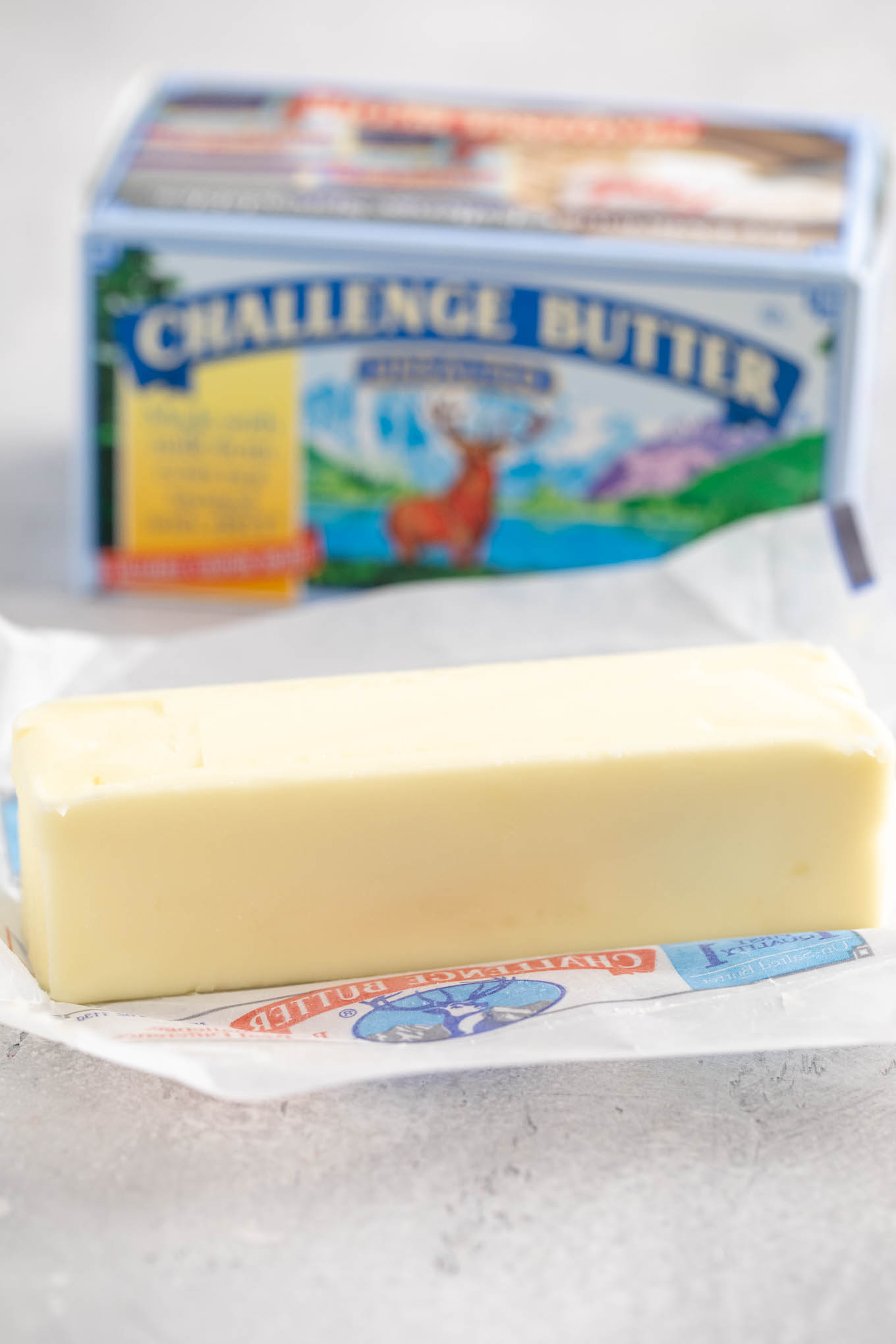 An opened stick of unsalted butter sitting on top of the wrapper. A box of butter is sitting in the background.