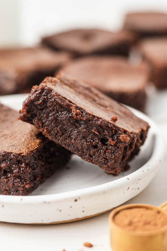 A close up view of two fudgy brownies on a white dessert plate, leaning against each other. 