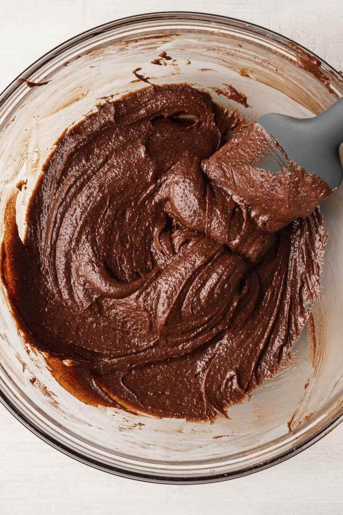 An overhead view of raw brownie batter in a glass mixing bowl with a rubber spatula.