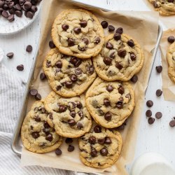 A pile of chocolate chip cookies on a baking sheet lined with parchment paper surrounded by more cookies and chocolate chips.