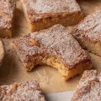 A snickerdoodle blondie with a bite taken out on a brown piece of parchment paper. More blondies are scattered around it.