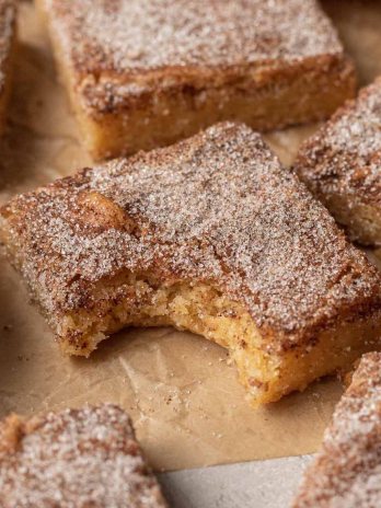 A snickerdoodle blondie with a bite taken out on a brown piece of parchment paper. More blondies are scattered around it.