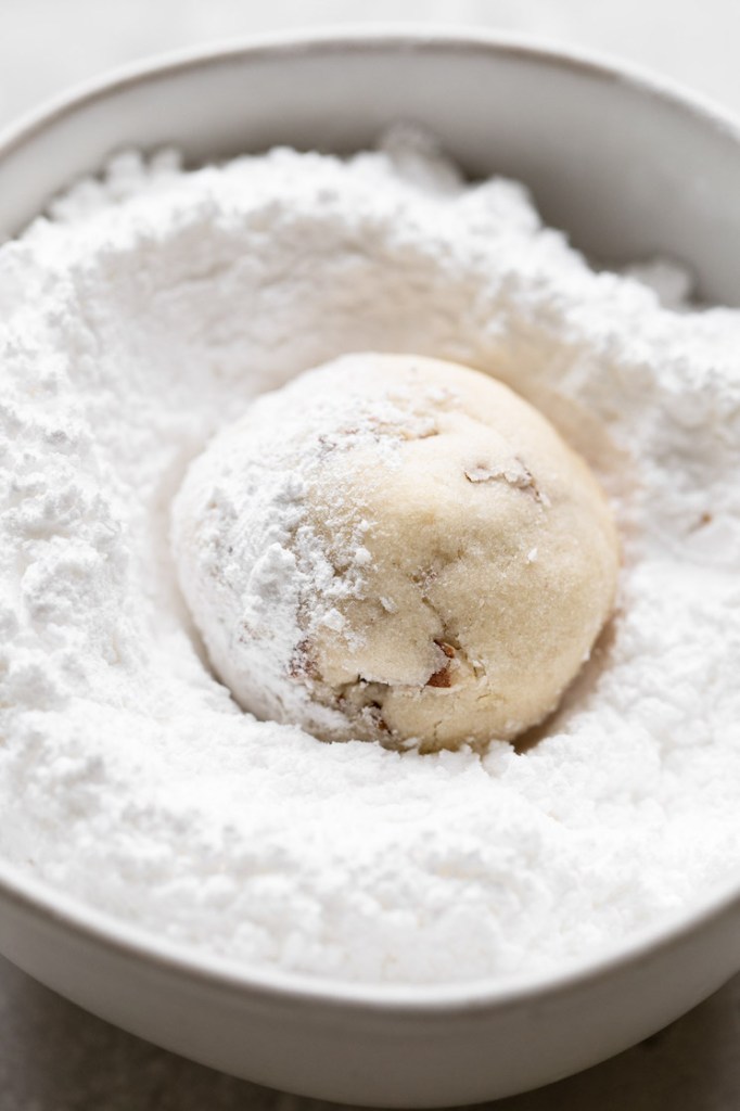 A baked snowball cookie being rolled in powered sugar before being set aside to cool completely.