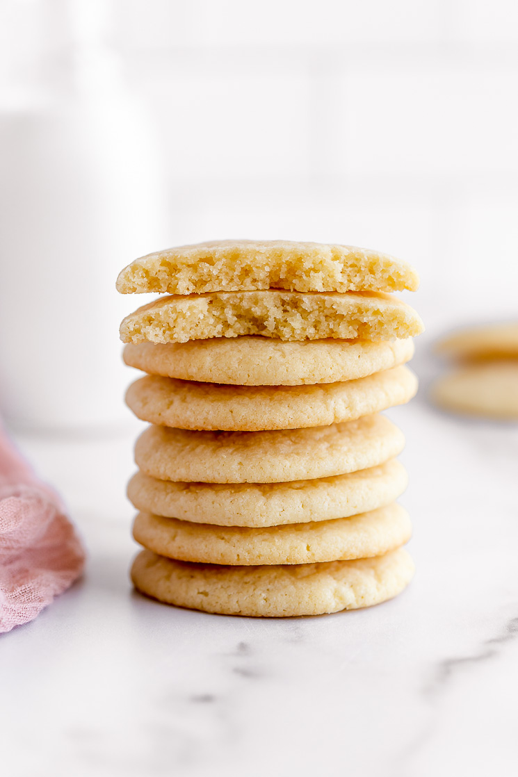 A stack of soft and chewy sugar cookies, resting on a marble surface, with the top cookie broken in half to show inside texture.