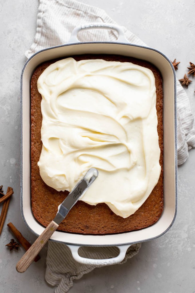 The cake being frosted with cream cheese frosting in a nine by thirteen pan.