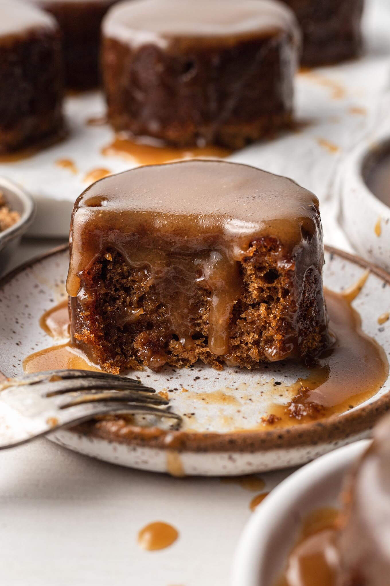 A piece of sticky toffee pudding on a white speckled plate. A bite has been taken out and a fork rests on the plate.