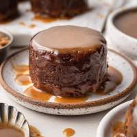 A small round date cake covered in toffee sauce on a white speckled plate.