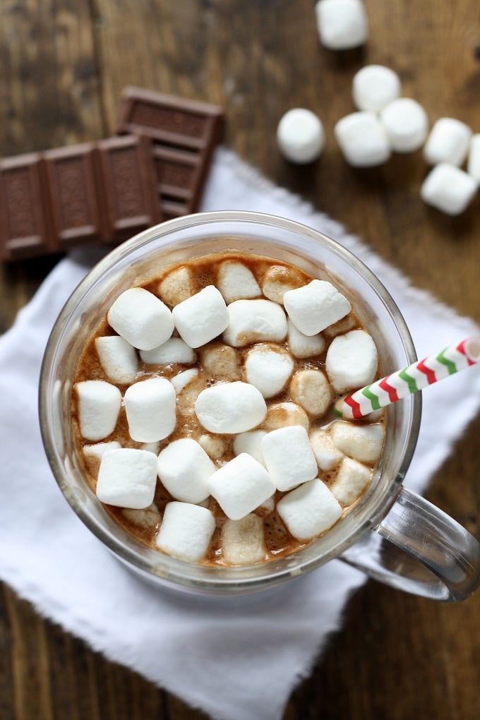 Overhead view of hot chocolate topped with marshmallows in a glass mug. Chocolate squares and mini marshmallows rest in the background. 