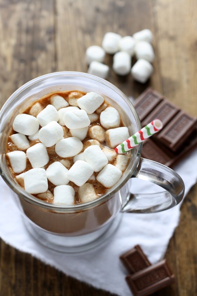 Mug of homemade hot chocolate topped with mini marshmallows and a straw. Chocolate squares and marshmallows rest in the background. 