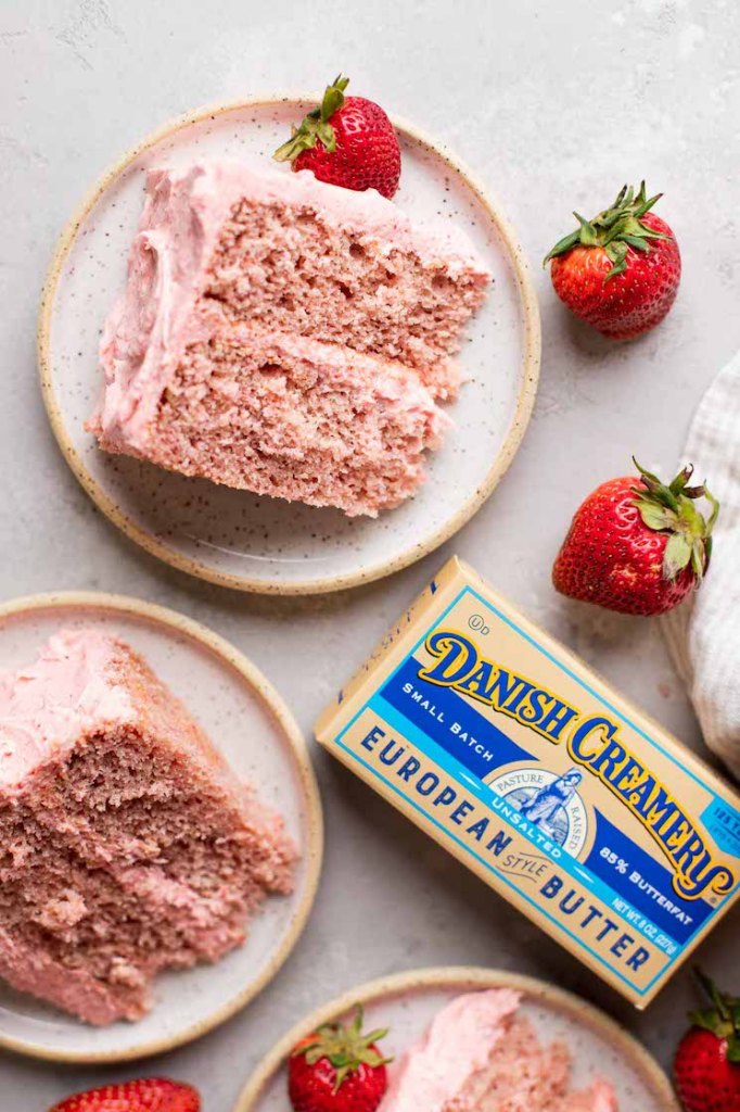 An overhead view of a few slices of strawberry cake on white speckled plates. A box of butter and strawberries surround the plates.