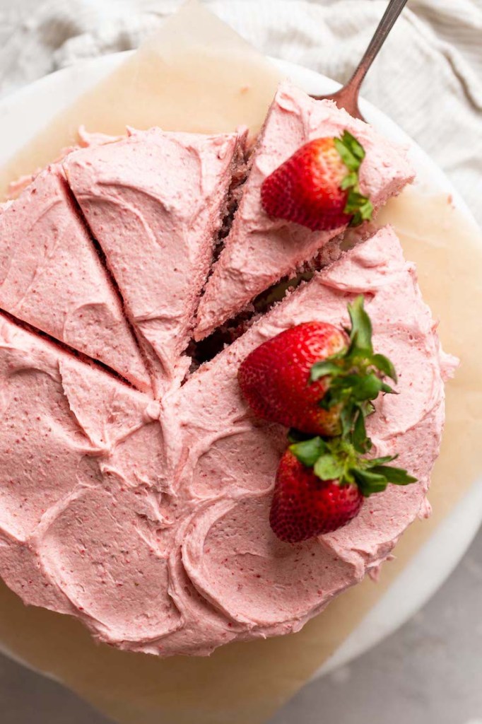 An overhead view of a strawberry cake. The cake has been sliced and one slice is being removed.