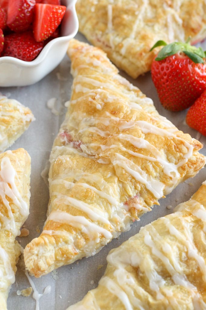 Several strawberry turnovers on a piece of parchment paper. Some sliced strawberries inside a white bowl sit next to the turnovers.