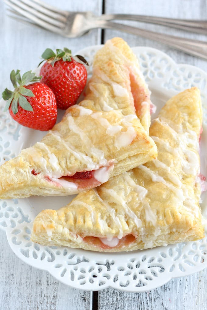 Two strawberry turnovers on a decorative white plate. Two strawberries rest on the plate and two forks rest in the background.