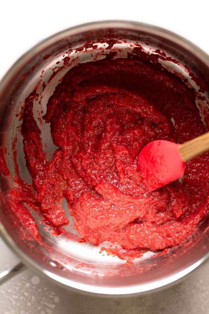 A saucepan filled with reduced strawberry puree. A rubber spatula rests on the side of the pan.