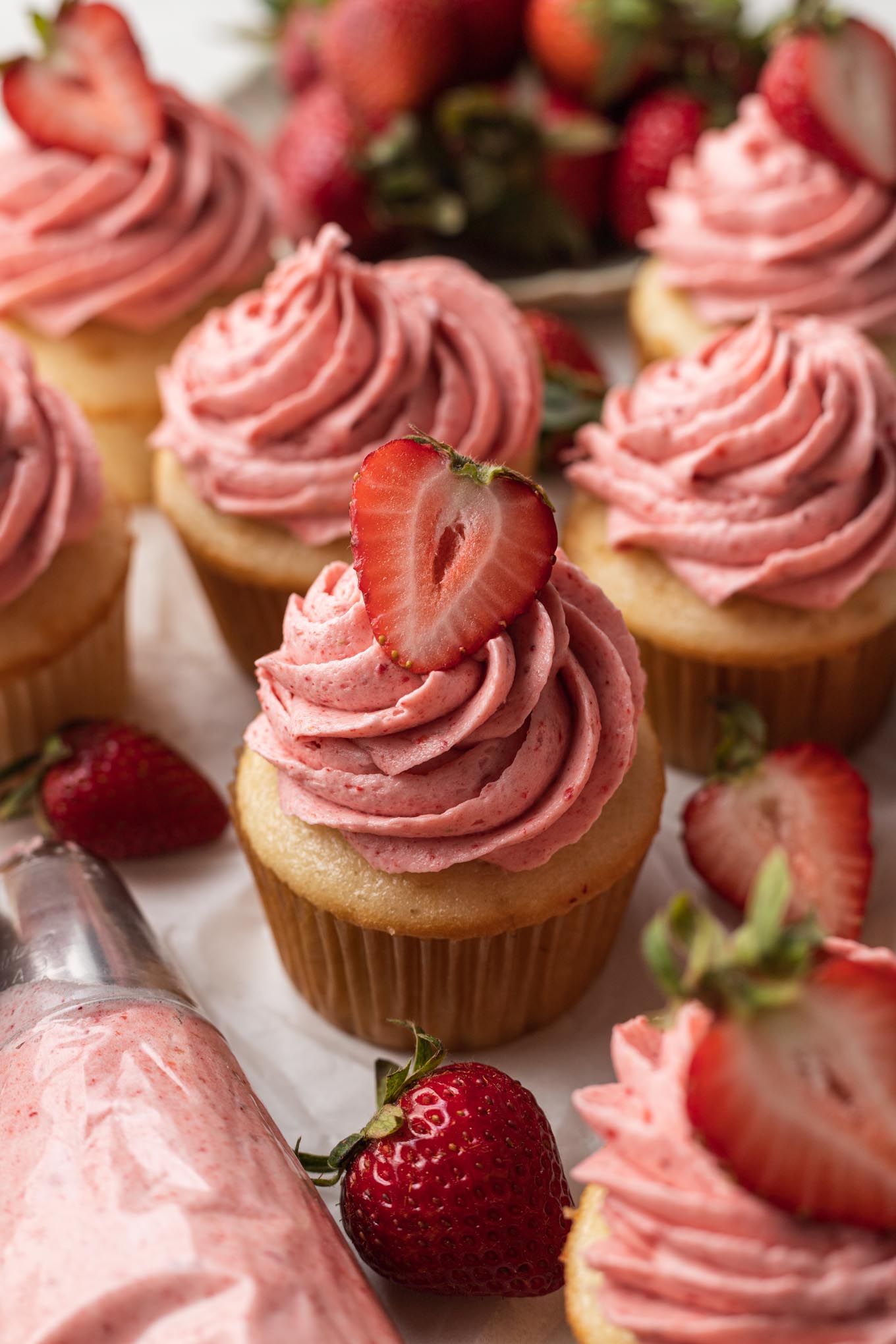 Several vanilla cupcakes topped with strawberry frosting. A piping bag with frosting rests near the cupcakes.