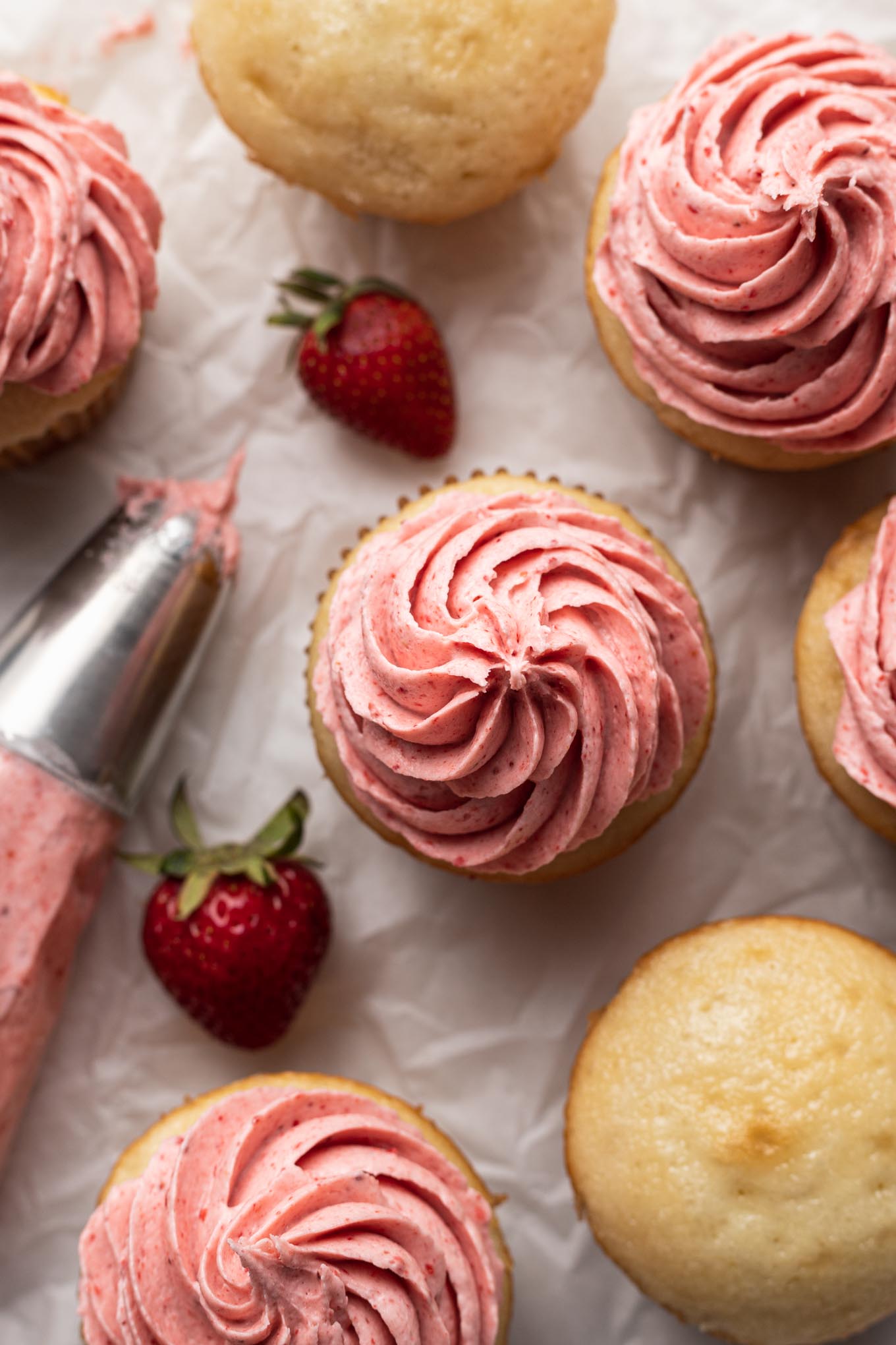 An overhead view of strawberry frosted cupcakes, fresh berries, and a piping bag. 