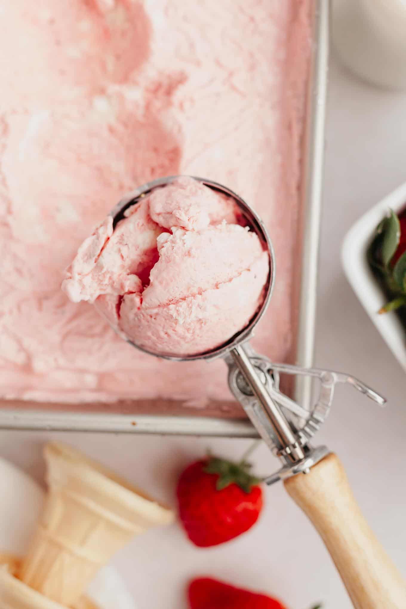 A scoop of strawberry ice cream held over a pan of ice cream. 