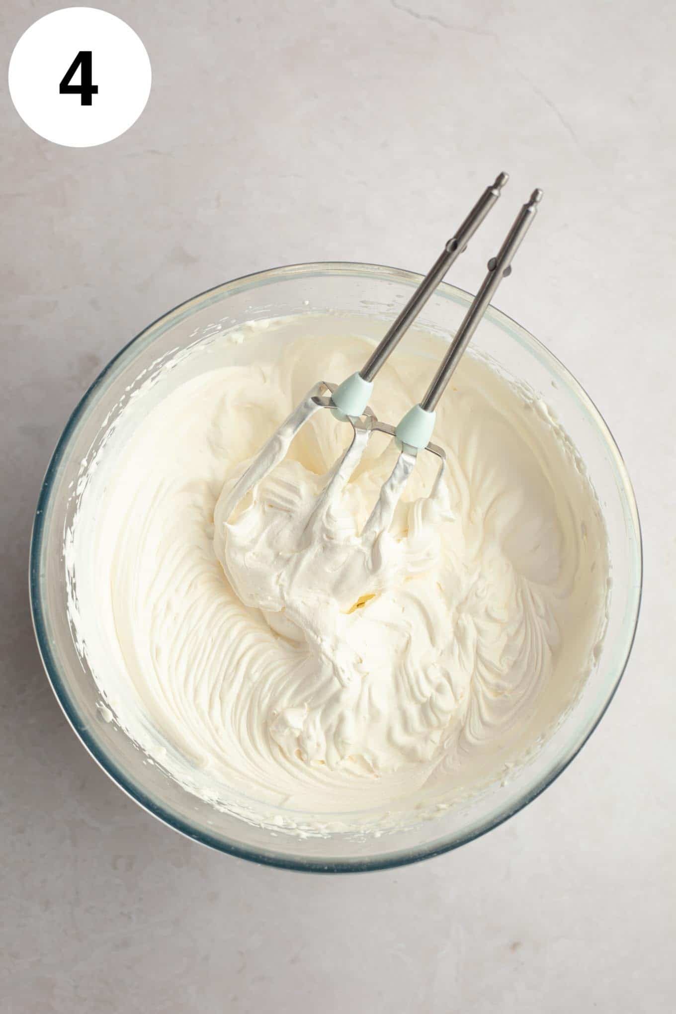 An overhead view of whipped cream in a glass mixing bowl, with two electric beaters. 