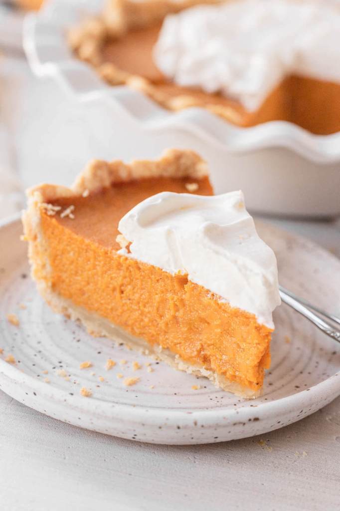 A slice of sweet potato pie topped with whipped cream on a white speckled dessert plate. The rest of the pie rests in the background. 