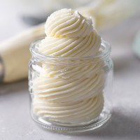 Swiss meringue buttercream frosting piped into a glass jar. A piping bag lies in the background.