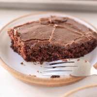 A slice of Texas sheet cake on a white speckled plate. A bite has been taken out of the slice and a fork rests on the side of the plate.