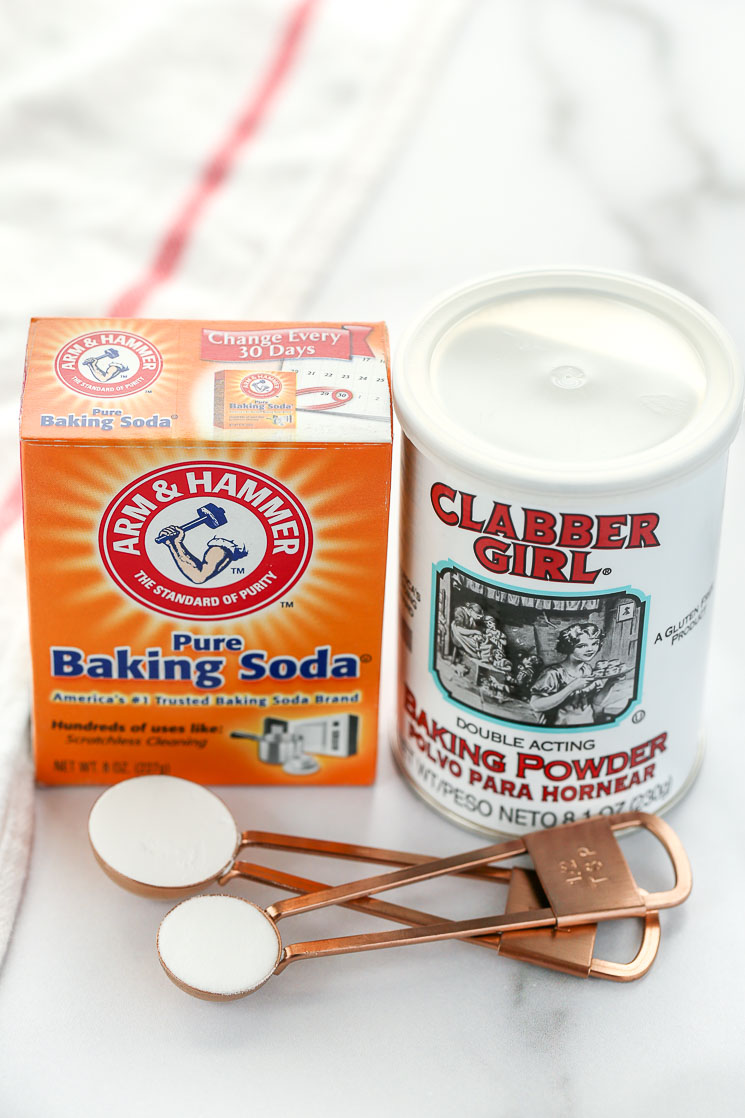 A box of baking soda, can of baking powder, and measuring spoons sitting on top of a marble surface with a white napkin on the side.