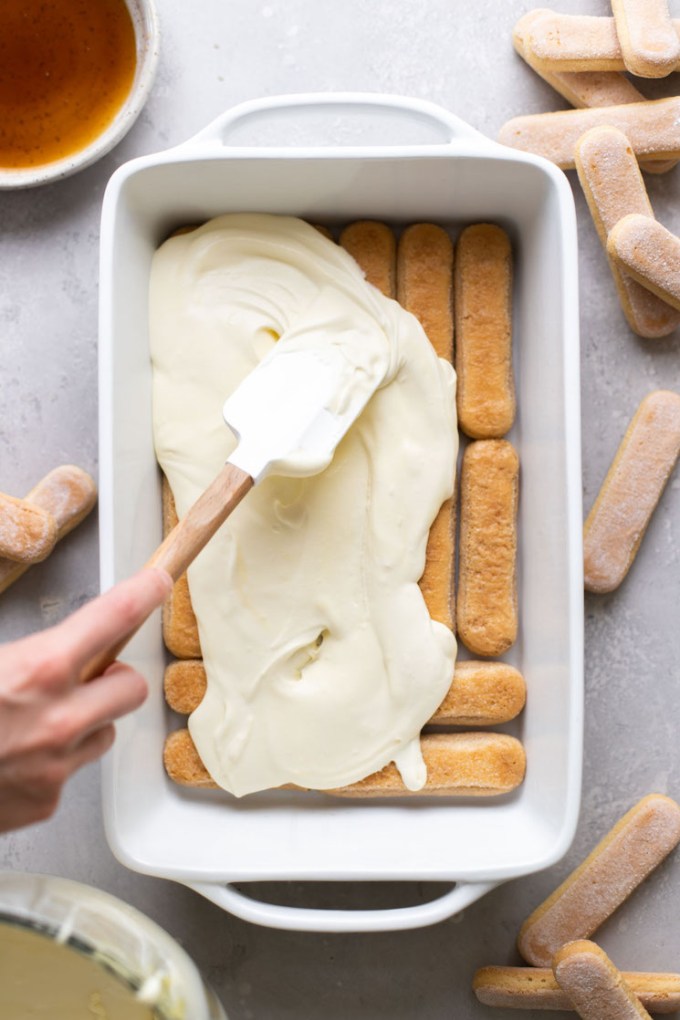 Overhead view showing mascarpone filling being spooned onto coffee-dipped lady fingers in a white baking dish.