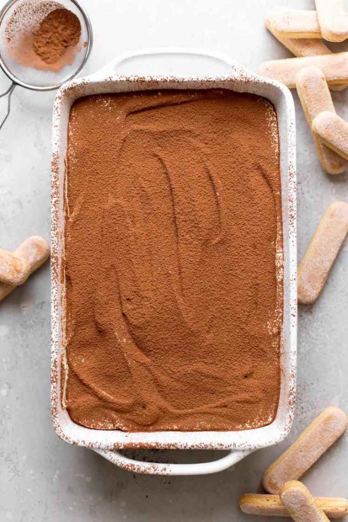 Overhead view of a baking dish of homemade tiramisu. Lady finger cookies are scattered around the dish.