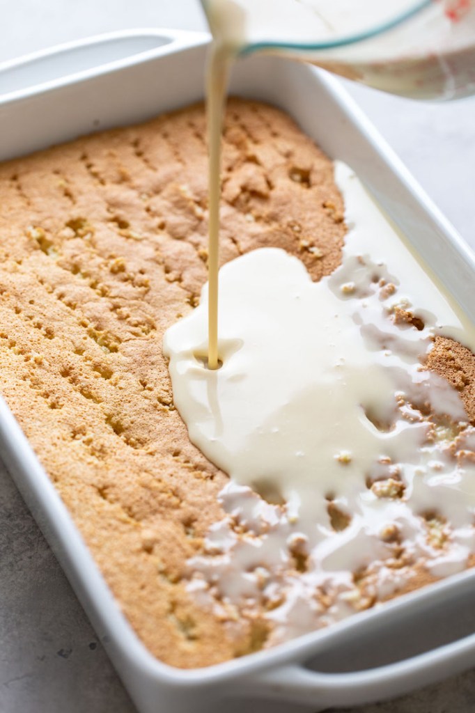 Three milk filling being poured over a sponge cake that's been poked all over with a fork. 