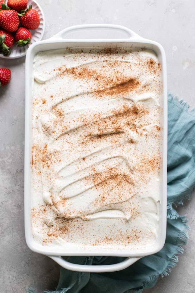 Overhead view of an authentic tres leches cake in a baking dish. A bowl of fresh strawberries rests nearby. 