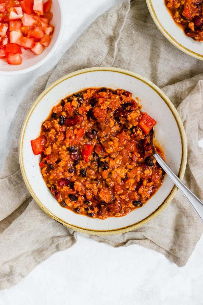 A bowl of turkey chili with a spoon resting in it. Another bowl of diced tomatoes and chili rest nearby.