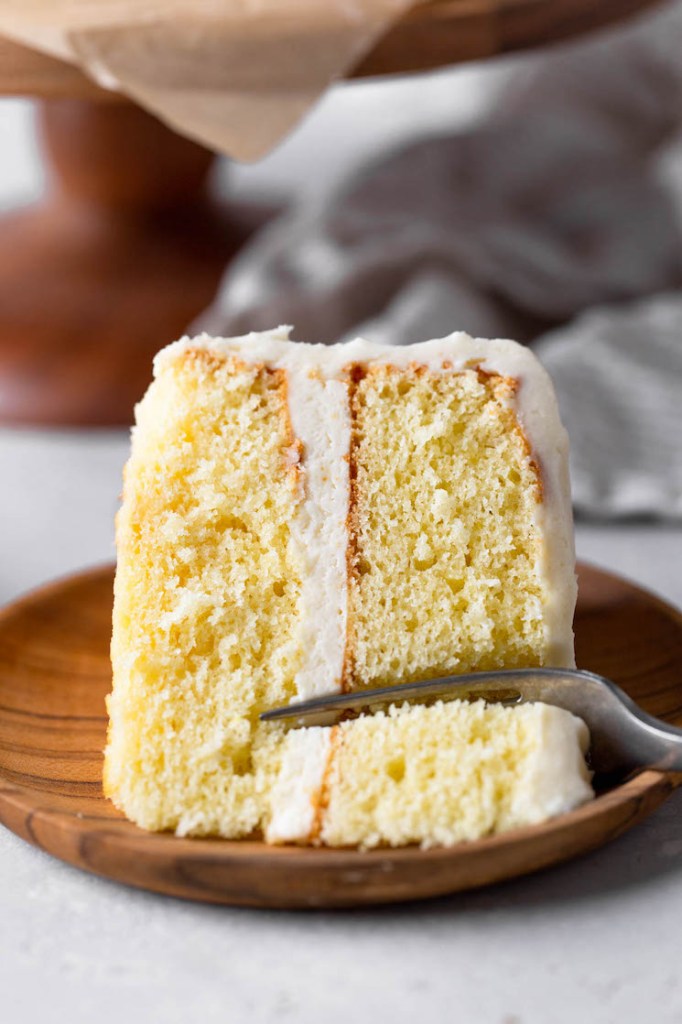 A slice of vanilla cake lying on its side on a wood plate. A fork is digging into the end of the slice. 