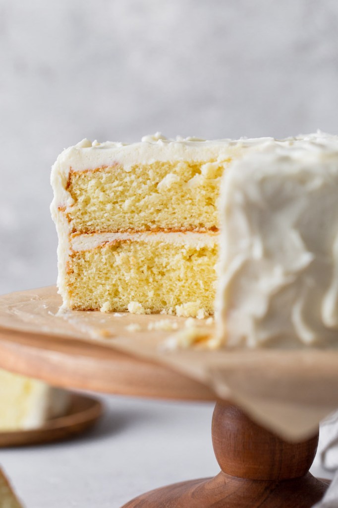 A fluffy vanilla cake frosted with vanilla buttercream, viewed from the side. A slice has already been removed from the cake stand. 