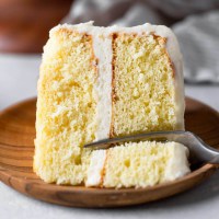 A slice of vanilla cake lying on its side on a wood plate. A fork is digging into the end of the slice.