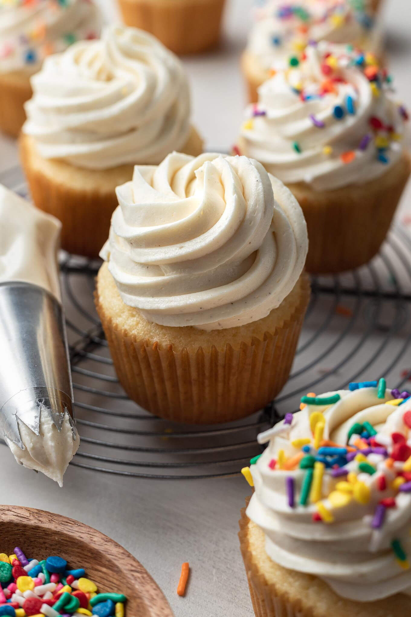 Frosted vanilla cupcakes next to a piping bag and dish of sprinkles. 