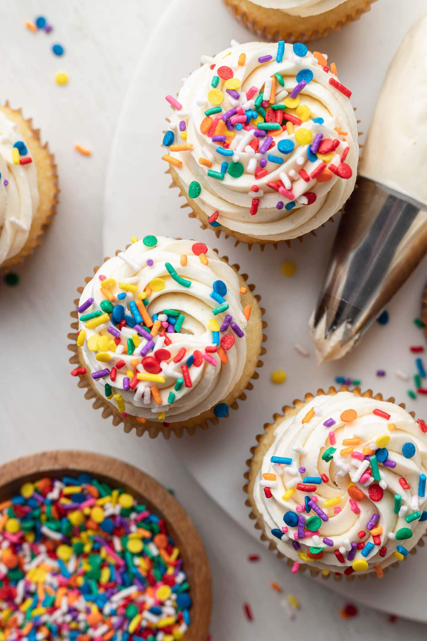 An overhead view of three cupcakes with white frosting next to a piping bag and sprinkles. 