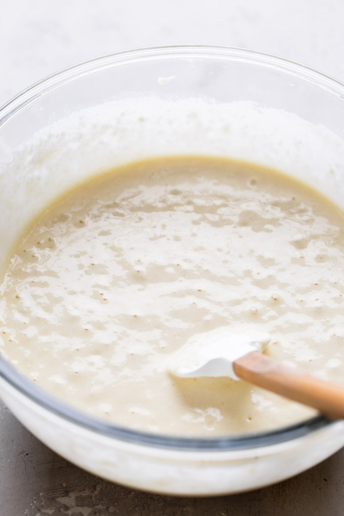 A glass mixing bowl full of waffle batter. A rubber spatula rests in the bowl.