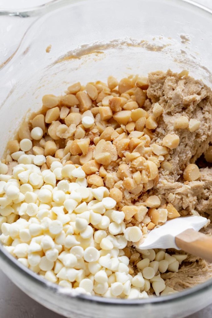 A glass mixing bowl filled with cookie dough topped with white chocolate chips and nuts being mixed together with a rubber spatula.