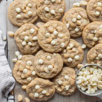 A batch of cookies laid out on a piece of parchment paper with extra nuts and white chocolate chips beside them.