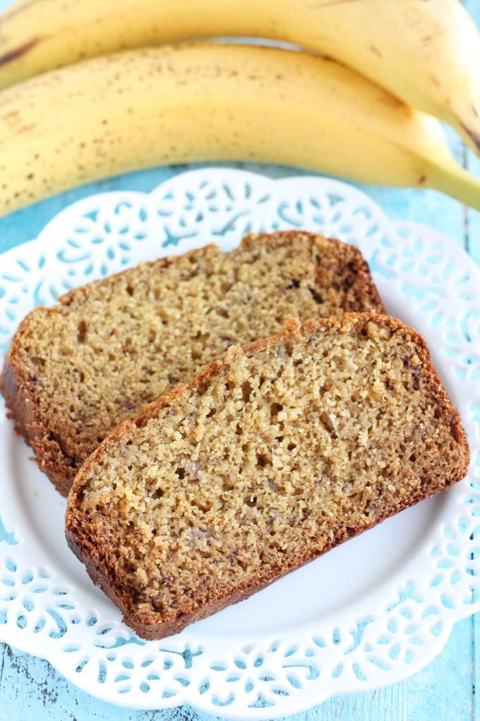 Two slices of banana bread with wheat flour on a white plate. Two bananas rest in the background. 