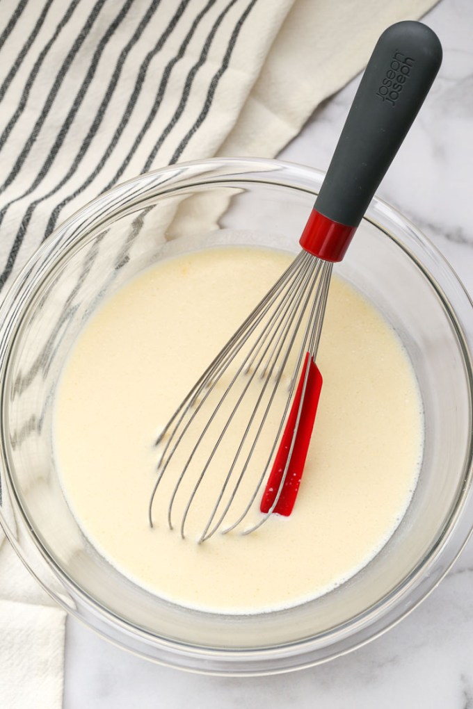 A clear bowl filled with the wet ingredients for pancake batter and a red and black whisk.