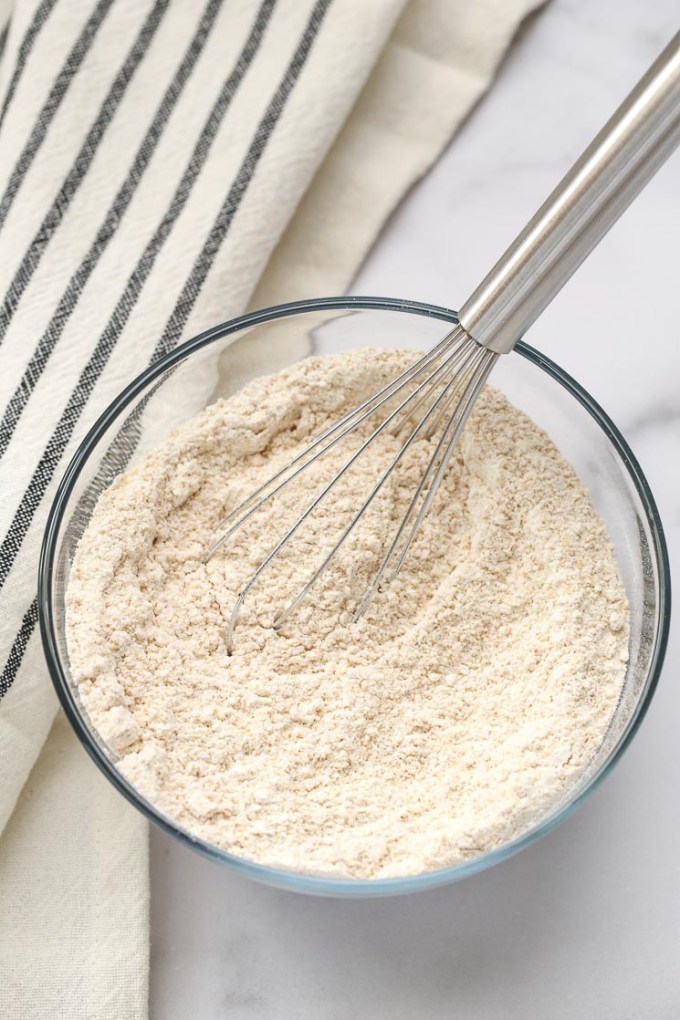 A clear bowl filled with white whole wheat flour, baking powder, baking soda, and salt with a silver whisk.