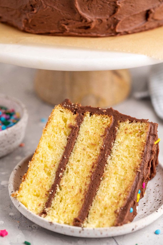 A slice of yellow cake on a white speckled plate. The rest of the cake is sitting on a cake stand in the background.