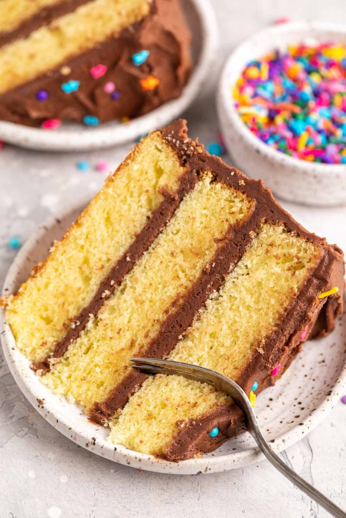 Two slices of yellow cake on white speckled plates. A fork is cutting into one slice of cake.