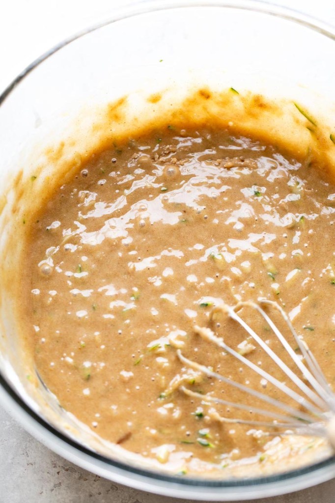 An overhead view of zucchini muffin batter in a glass mixing bowl with a whisk. 