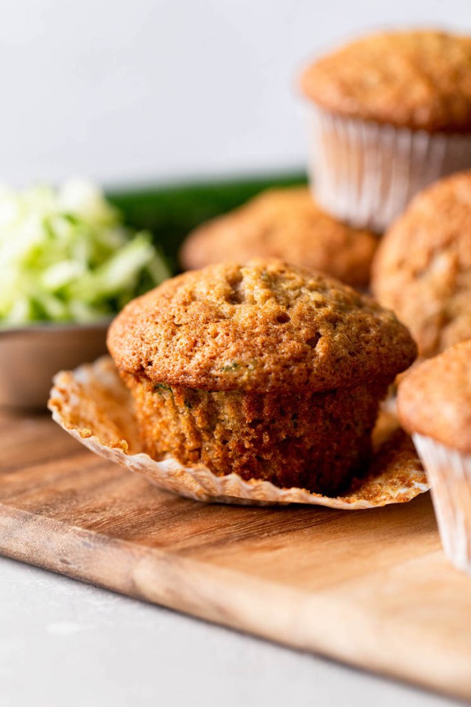 A close up of a zucchini muffin with the muffin liner pulled down. It's on a wood cutting board with more muffins surrounding it. 